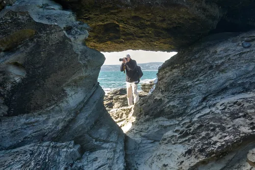 Rocky photographer The image is a landscape photograph taken on the coast during a late morning in early autumn. The main subject is a person known as Rocky Photographer, who is standing on sunlit rocks beneath a natural rock arch, facing the ocean while holding a camera to take photographs. The sea and distant coastline are visible in the background with gentle waves, and the weather appears clear with a bright sky. Michael Weinbren captured the scene, which highlights the rugged coastal environment and the activity of photography in a natural setting.